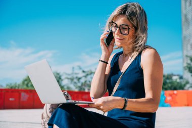 Donanma elbiseli bir kadın telefonuyla konuşurken gülümsüyor ve laptopuyla çalışıyor, açık havada şehirde oturuyor..