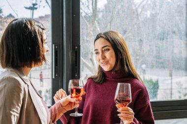 Pencere kenarında duran iki kadın gül şarabı bardağını tutuyor. Gülümsüyorlar ve sohbet ediyorlar, birlikte rahat bir anın tadını çıkarıyorlar..