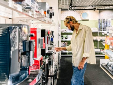 Bir adam perakende mağazasında çeşitli kahve makinelerine göz atar, farklı modellerin özelliklerini ve tasarımlarını dikkatle inceler....