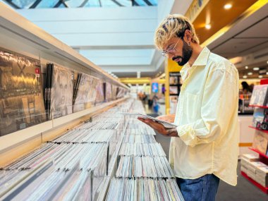 Gözlüklü ve açık renk gömlekli bir adam modern bir mağazada plak plaklarını karıştırıyor. Koleksiyondaki diğerlerini kontrol ederken bir kayıt tutuyor....