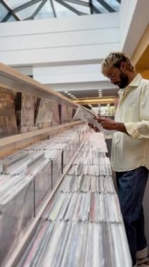 Gözlüklü ve açık renk gömlekli bir adam modern bir mağazada plak plaklarını karıştırıyor. Koleksiyondaki diğerlerini kontrol ederken bir kayıt tutuyor..