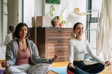 Yoga minderleri üzerinde bağdaş kurmuş, rahat bir odada bitki ve güneş ışığıyla meditasyon yapan iki kadın. Huzurlu ve odaklı ifadeler.