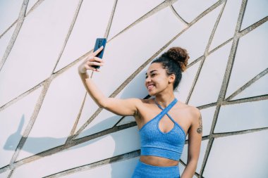 Mavi aktif giysili fit bir kadın modern geometrik bir duvarın önünde selfie çekerken gülümsüyor ve egzersiz anını yakalıyor..