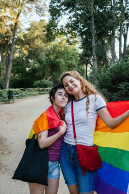 Birbirini kucaklayan iki kadın parkta yürürken omuzlarında gökkuşağı bayrağı taşıyorlar.