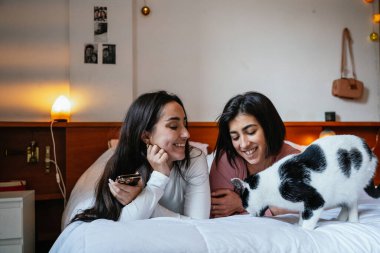 İki kadın, siyah beyaz kedilerini yatakta yürürken seyrederken sıcak bir şekilde gülümsüyor, evcil hayvanlarıyla huzurlu bir anın tadını çıkarıyorlar..