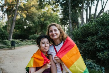 Parkta gökkuşağı bayrağı taşıyan iki kadın birbirini kucaklıyor.