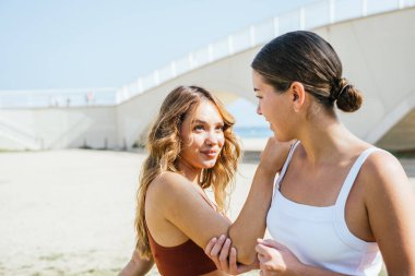 İki bayan arkadaş, antrenmandan sonra kumsalda birlikte esniyorlar, güneşin tadını çıkarıyorlar ve birbirlerine eşlik ediyorlar.