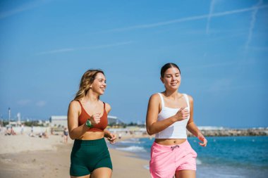 İki bayan sporcu sahilde koşuyor, deniz kenarında antrenmanlarının keyfini çıkarıyorlar.