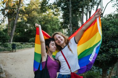 İki kadın kollarını açarak gökkuşağı bayrağını tutarken birbirlerine sarılıyorlar.