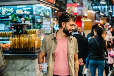 Elinde içecek tutan bir turist yürür ve Madrid, İspanya 'daki Mercado de San Miguel' deki yiyecek tezgahlarına bakar.