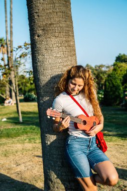 Parkta gitar çalan kıvırcık saçlı kadın palmiye ağacına yaslanıyor.