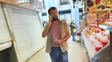 Elinde içecek tutan ve markette yürürken cep telefonuyla konuşan genç bir adam.