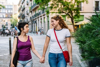 Güneşli bir yaz gününde iki genç kadın el ele tutuşuyor ve bir şehir caddesinde konuşuyor.