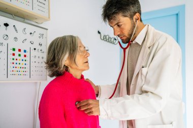 Steteskop kullanan bir erkek doktor klinikte tıbbi danışmanlık sırasında yaşlı bir kadın hastayı muayene ediyor.