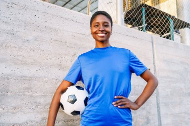 Beton duvarda futbol topuyla mavi gömlekli gülümseyen bir sporcu.