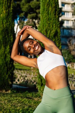 Gülümseyen sporcu kadın yeşil ağaçlar ve çimlerle kaplı bir parkta kollarını esnetiyor, beyaz bluz ve yeşil taytlar giyiyor.