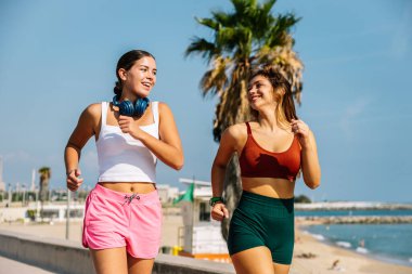 Güneşli bir günde kumsalda koşan iki bayan sporcu antrenmanlarının keyfini çıkarıyorlar.