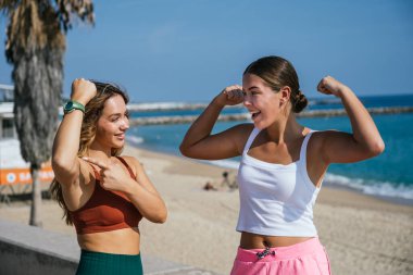 İki sağlıklı genç kadın güneşli bir sahilde kaslarını esnetiyor, güçlerini ve sağlıklı yaşam tarzlarını kutluyorlar.