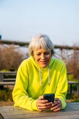 Parkta akıllı telefon kullanarak gülümseyen, emekliliğin tadını çıkaran ve bağlantıda kalan son sınıf öğrencisi.