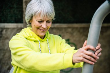 Mutlu olgun kadın ısınıyor ve şehir spor aletleri üzerinde esneme egzersizleri yapıyor.