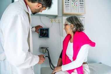 Erkek doktor, doktor muayenehanesinde kıdemli kadın hastanın kan basıncını ölçmek için sphygmomanometre kullanıyor.