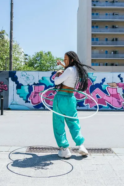 Örgülü genç bir kadın hula çemberleriyle şehir ortamında grafiti duvarlı, güneşli bir günün tadını çıkarıyor.