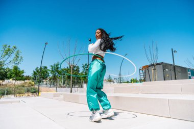 Genç bir kadın modern bir şehir parkında hula hop dansı yapıyor, yaz güneşinin ve temiz havanın tadını çıkarıyor.