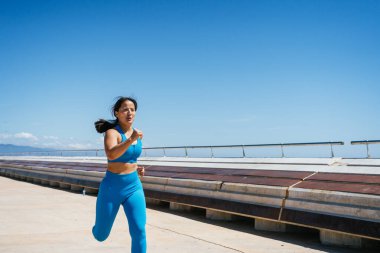 Atletik kadın açık mavi gökyüzünün altında kıyı yolunda koşuyor.