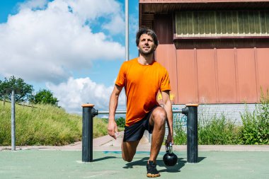 Erkek sporcu açık hava jimnastik parkında kettlebell ile hamleler yapıyor.