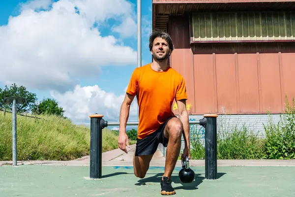 Erkek sporcu açık hava jimnastik parkında kettlebell ile hamleler yapıyor.