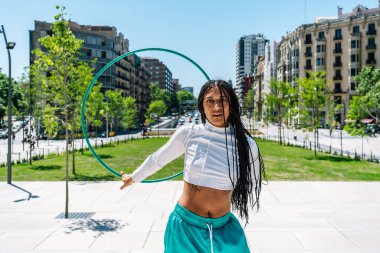 Uzun örgülü genç bir kadın şehir parkında hula çemberiyle dans ediyor, yaz havasının tadını çıkarıyor ve sağlıklı bir yaşam tarzının reklamını yapıyor.