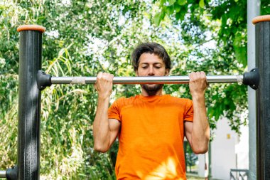 Kararlı erkek sporcu yemyeşil parktaki açık hava egzersiz ekipmanlarında barfiks çekiyor.