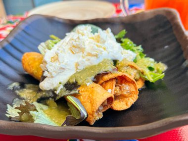 Salsa verde, krema ve cotija peyniri ile kaplı gevrek sarılmış tacolar, siyah bir tabakta servis edilir ve lezzetli Meksika mutfağı sergilenir.