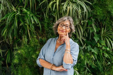 Confident senior woman posing against a green living wall, smiling and feeling happy