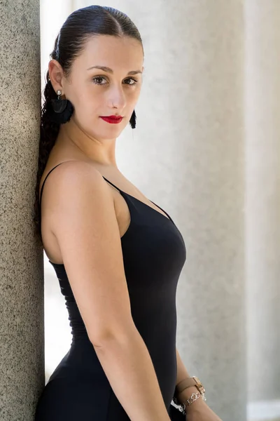 Vertical shot of a curly-haired young brunette woman leaning against a gray stone column wearing a black sundress with straps and a nose piercing