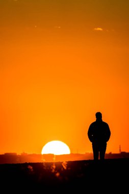 Silhouette of a man with a coat and his hands in his pockets, watching the sunset with buildings, buildings and a big sun on the horizon.