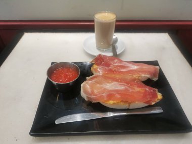 Sütlü kahve ve domatesli iki dilim tost ve siyah tabakta serrano jambonu ve domatesi yaymak için bir bıçakla kahvaltı..