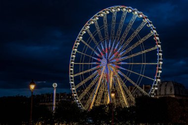 Fransız bayrağının renkleriyle aydınlatılmış bir dönme dolabın yatay gece görüntüsü. Kırmızı, beyaz ve mavi, bulutlu bir gökyüzü ve arka planda Paris 'in tipik çatıları..