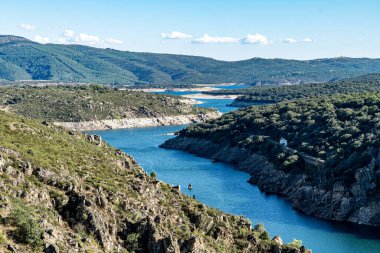 Alçak bir dağ bölgesindeki doğa manzarasında gezinen bir nehir yatağı tasviri. Açık mavi gökyüzü, çalılar ve çok yeşil bitki örtüsüyle dolu..