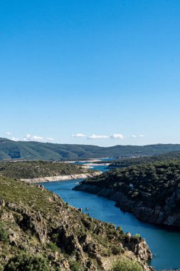 Alçak bir dağ bölgesindeki doğa manzarasında gezinen bir nehir yatağı tasviri. Açık mavi gökyüzü, çalılar ve çok yeşil bitki örtüsüyle dolu..