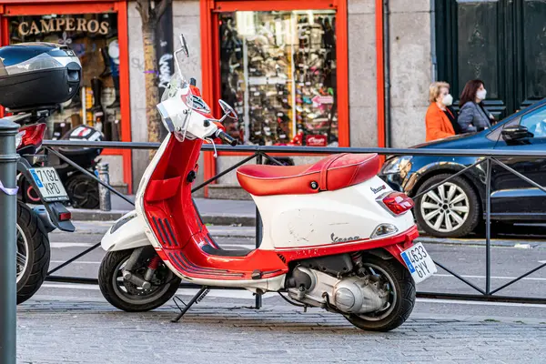 Madrid, Espaa. 6 de abril de 2021. Kırmızı-beyaz Vespa scooter 'ı Madrid' in merkezinde işlek bir caddenin kenarındaki kaldırıma park etmiş, arka planda vitrinler ve yayalar var..
