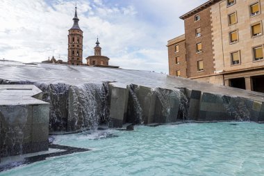 Zaragoza, İspanya. 20 Nisan 2025. Su Zaragozas Plaza del Pilar 'daki Fuente de la Hispanidad' ın üzerinden akıyor. Tarihi kilise kuleleri ve binaları parlak bulutlu gökyüzü altında..