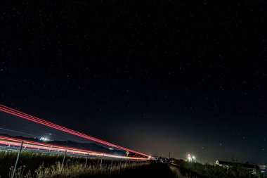 Yıldızlarla dolu bir gökyüzü altında kırmızı ve beyaz ışık yollarının bulunduğu bir İspanyol otoyolunun uzun gece fotoğrafı. Şehir hareketlerini evrenin sakinliğiyle harmanlıyor..