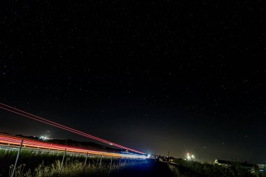 Yıldızlarla dolu bir gökyüzü altında kırmızı ve beyaz ışık yollarının bulunduğu bir İspanyol otoyolunun uzun gece fotoğrafı. Şehir hareketlerini evrenin sakinliğiyle harmanlıyor..
