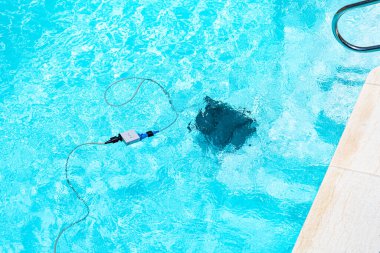 Temiz mavi bir havuzun havuzu ve hareket halindeki bir robot temizleyicisi. Beyaz fayanslarla, yeşil sentetik çimlerle ve güneşli bir günde metal merdiven ile çevrili..