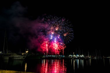 Uzun süren havai fişek gösterileri geceyi bir limanın üzerinde aydınlatıyor. Denizde renkli yansımalar, demirli yatlar, direklerin siluetleri ve siyah gökyüzünde kopyalama alanı var..