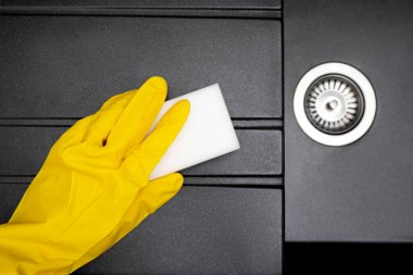 Sarı eldivenli bir kadının eli lavaboyu melamin süngerle yıkıyor. Yeni granit mutfak lavabosu, siyah yıkama düzlüğü. Ev temizleme servisi. Üst görünüm.