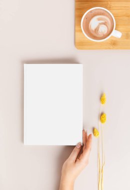 Bej bir arka planda, kahve ve kurutulmuş falaris çiçeğiyle bir kadın eli tarafından tutulan beyaz ciltli kitabın bir kopyası. İskandinav ya da wabi sabi tarzı tasarım ve reklamlarınız için boş şablon