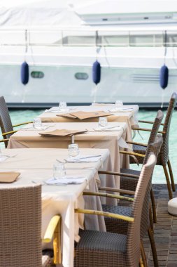 Arka planda yatları olan bir marina tarafından bardaklar ve peçetelerle hazırlanmış açık hava yemek masaları lüks bir yemek atmosferi yaratıyor. Seyahat ve konukseverlik temaları için ideal