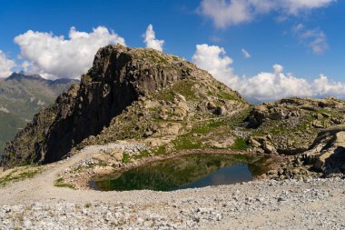 Serene small alpine lake with clear reflection of rocky cliffs in a high altitude rugged mountain landscape, surrounded by pristine nature and blue skies clipart